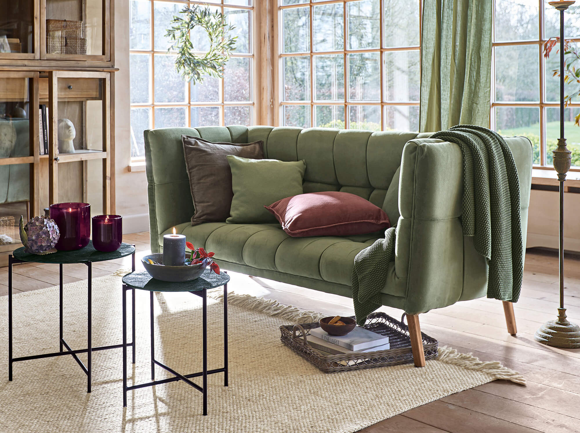 Groene fluwelen vintage bank met kussens en deken in lichte woonkamer met grote ramen