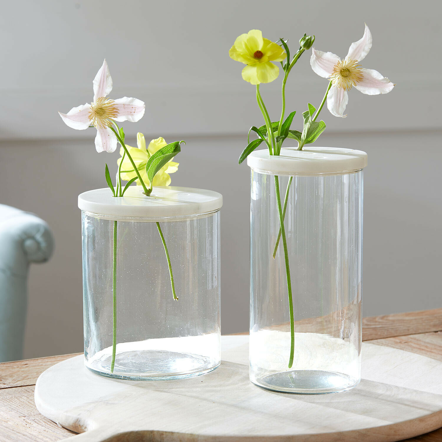 Glazen pot set van 2 Rosanella Twee transparante glazen vazen met witte deksels en lichte bloemen op een houten tafel