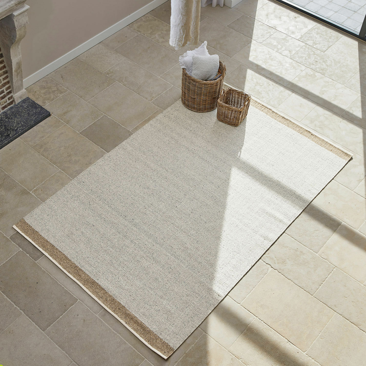 Beige vloerkleed van natuurlijk materiaal met bruine rand op lichte stenen vloer, interieurdecoratie