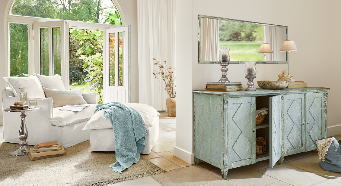 Gezellige witte fauteuil met voetenbank en blauwe deken naast groene vintage dressoir in lichte woonkamer