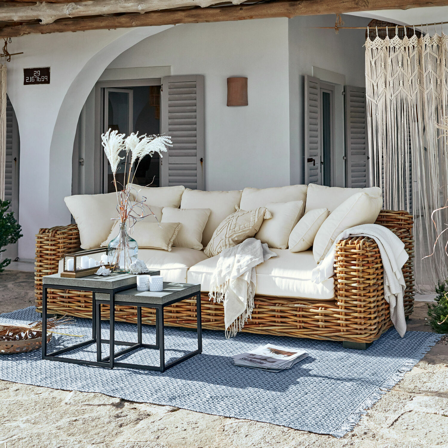 Bank Rimini Rotan bank met crèmekleurige kussens en deken op blauwe vloerkleed op zonnig terras