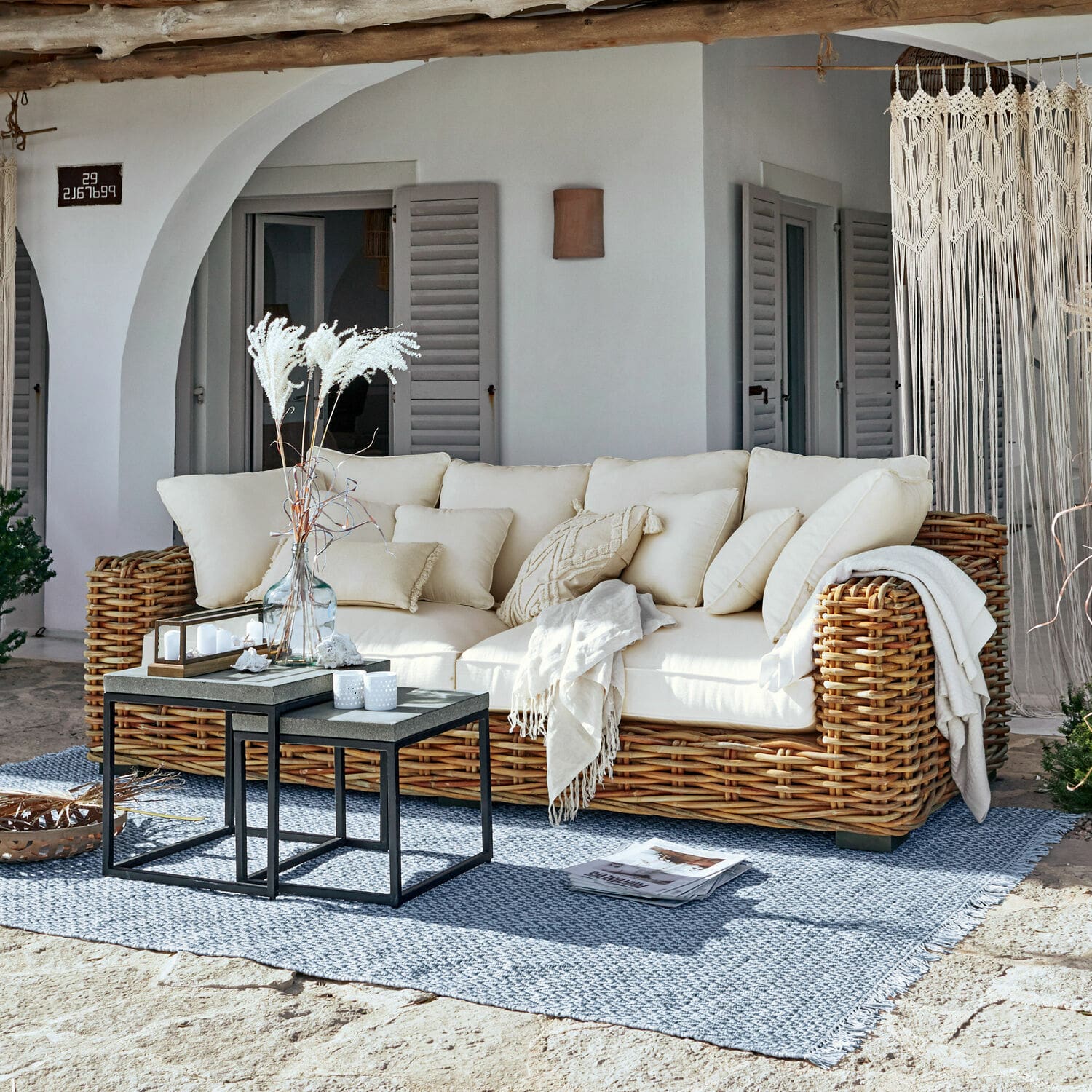Bank Rimini Rotan bank met crèmekleurige kussens en deken op blauwe vloerkleed op zonnig terras