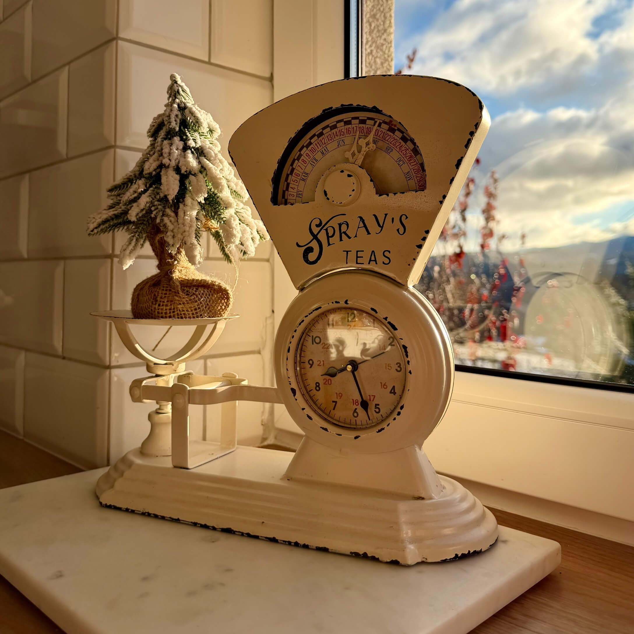 Beige metalen keukenweegschaal met klok en kerstdecoratie op vensterbank bij raam