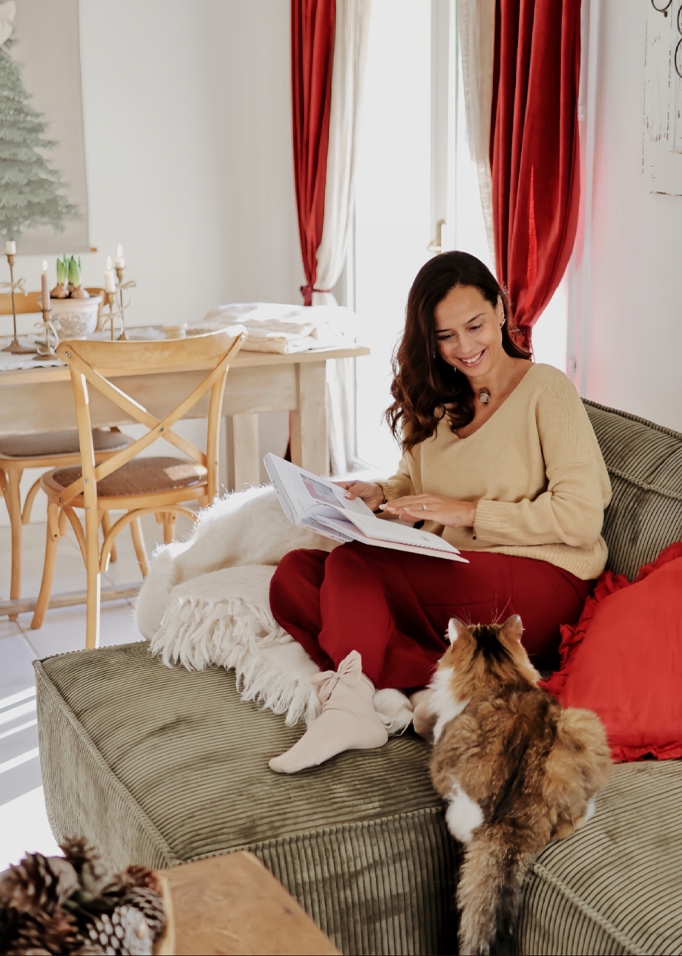 Vrouw in beige trui en rode broek leest boek op groene bank met kat in gezellige woonkamer