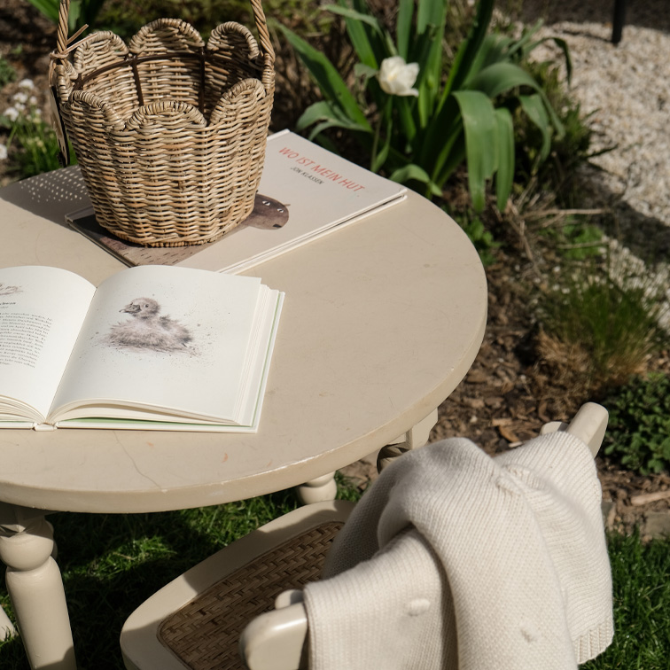 Ronde beige tuintafel met een rieten mand en open boeken op het gras in de zon