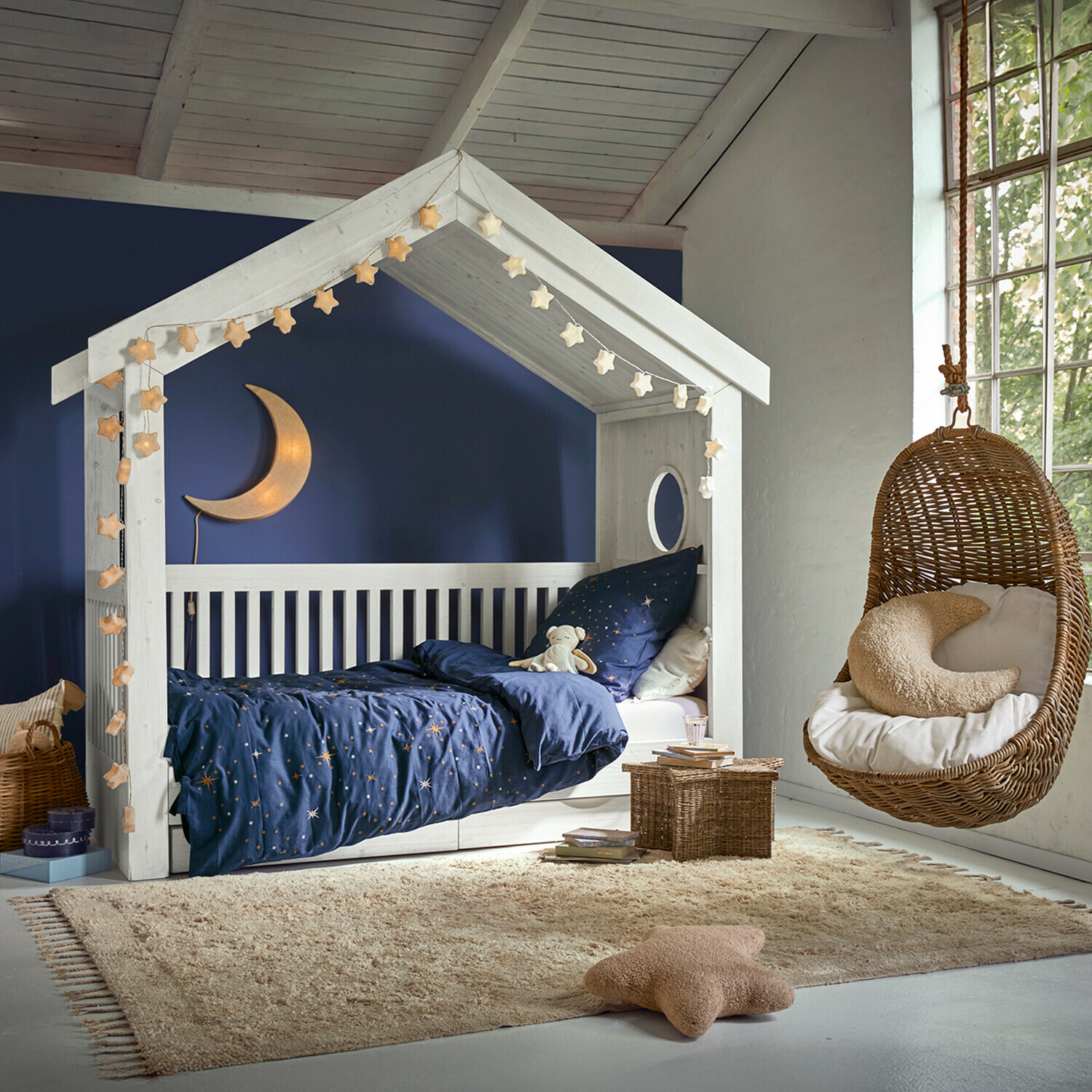 Wit houten huisvormig bed met donkerblauw sterrenbeddengoed en hangende rotan stoel in kinderkamer