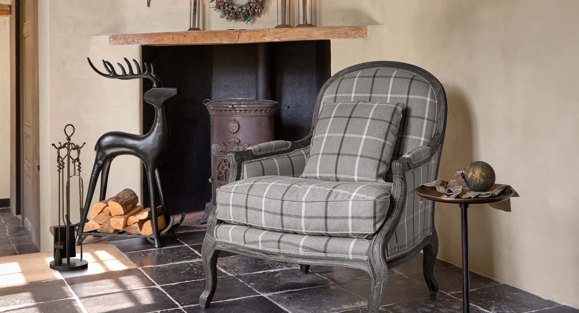 Grijze geruite fauteuil met houten frame naast open haard met metalen houtrek en gereedschap