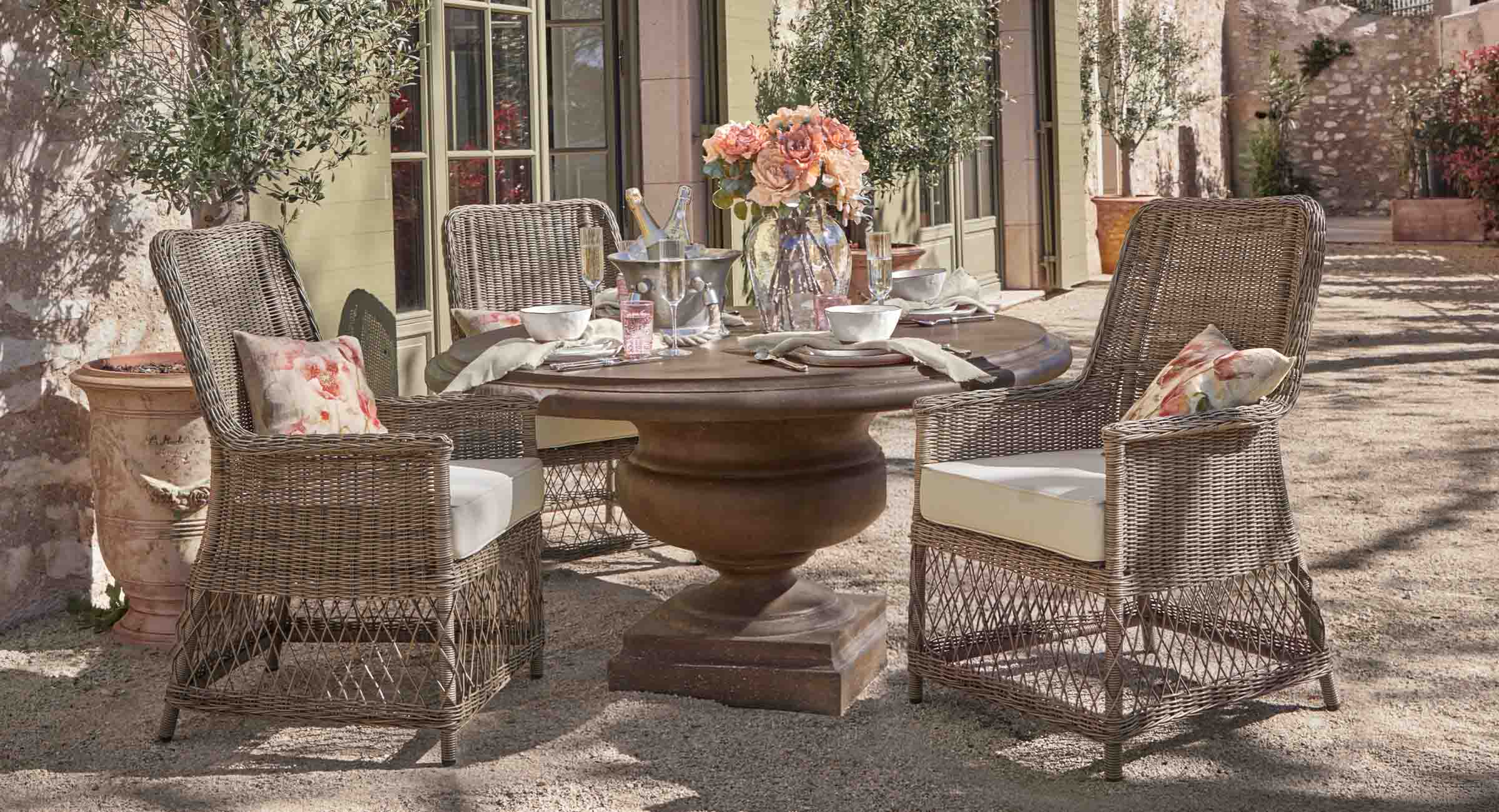 Ronde houten tuintafel in bruin met vier rotan stoelen met lichte kussens en bloemen als decoratie