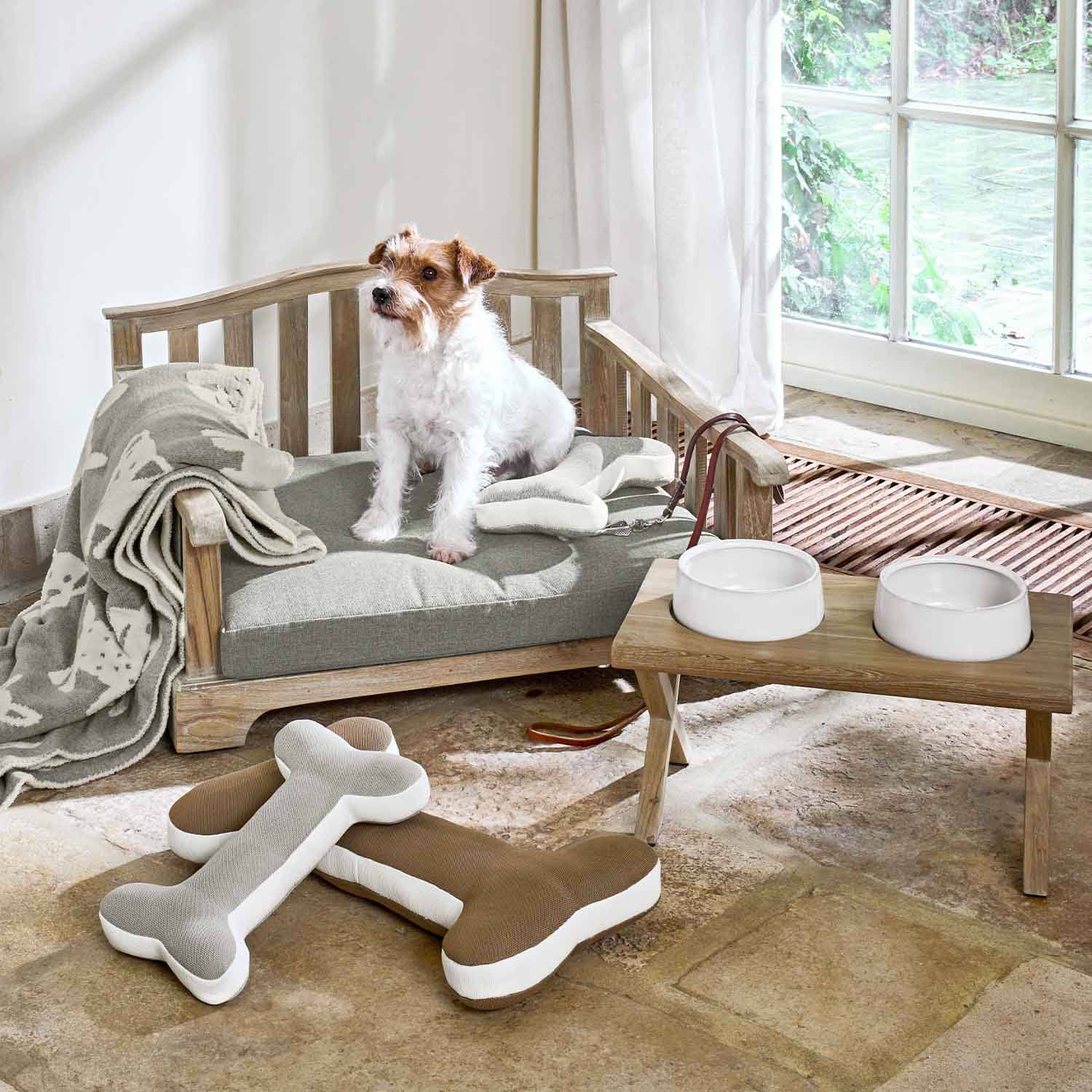 Houten hondenbed met grijs kussen, pluchen botten en witte voerbakken op houten standaard in kamer
