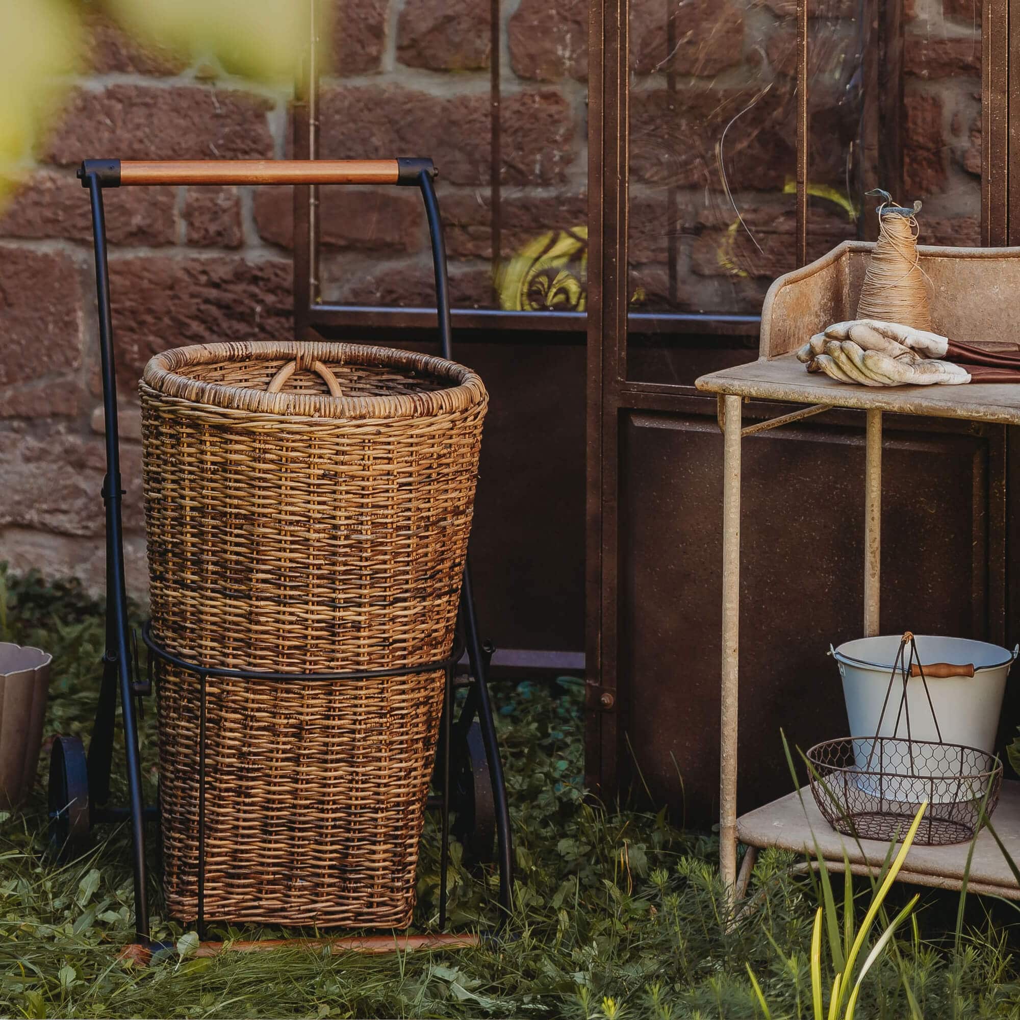 Wagen met rieten mand en metalen frame op gras bij bakstenen muur, tuingereedschap ernaast