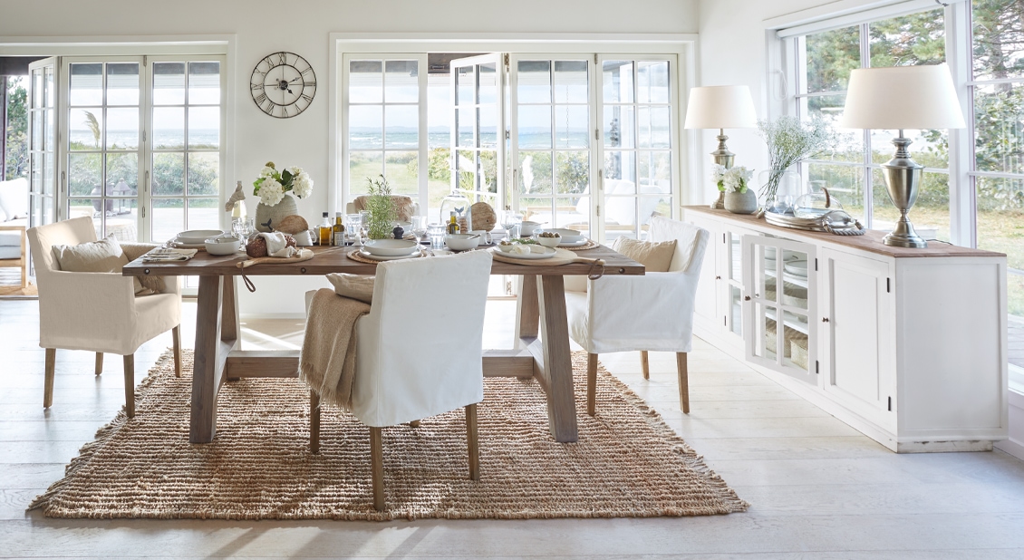Eetkamer in kuststijl met houten tafel en witte stoelen op natuurlijke vloerkleed met uitzicht op tuin