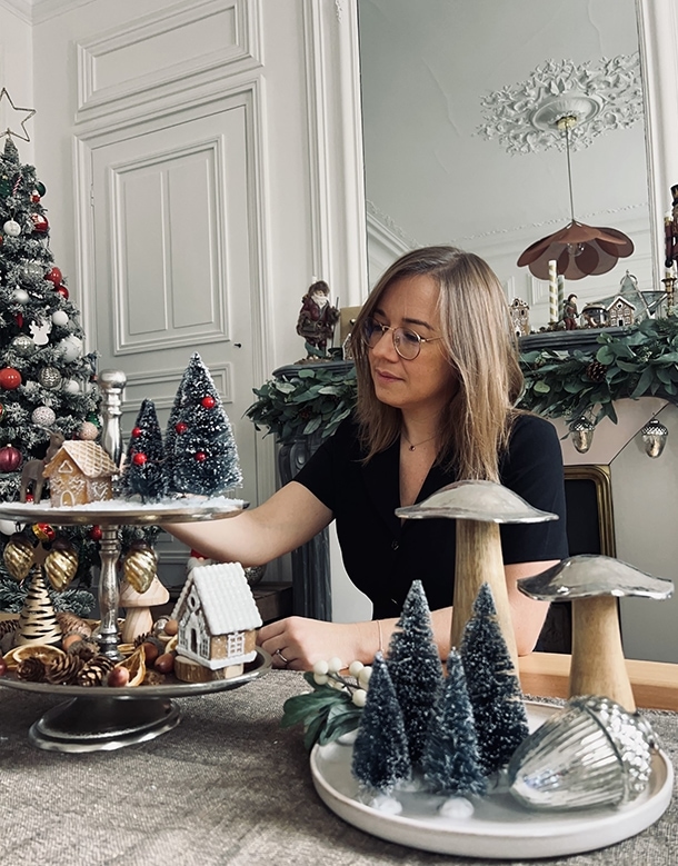 Vrouw die kersttafel versiert met miniatuurdennenbomen en huisjes in een gezellige kamer.