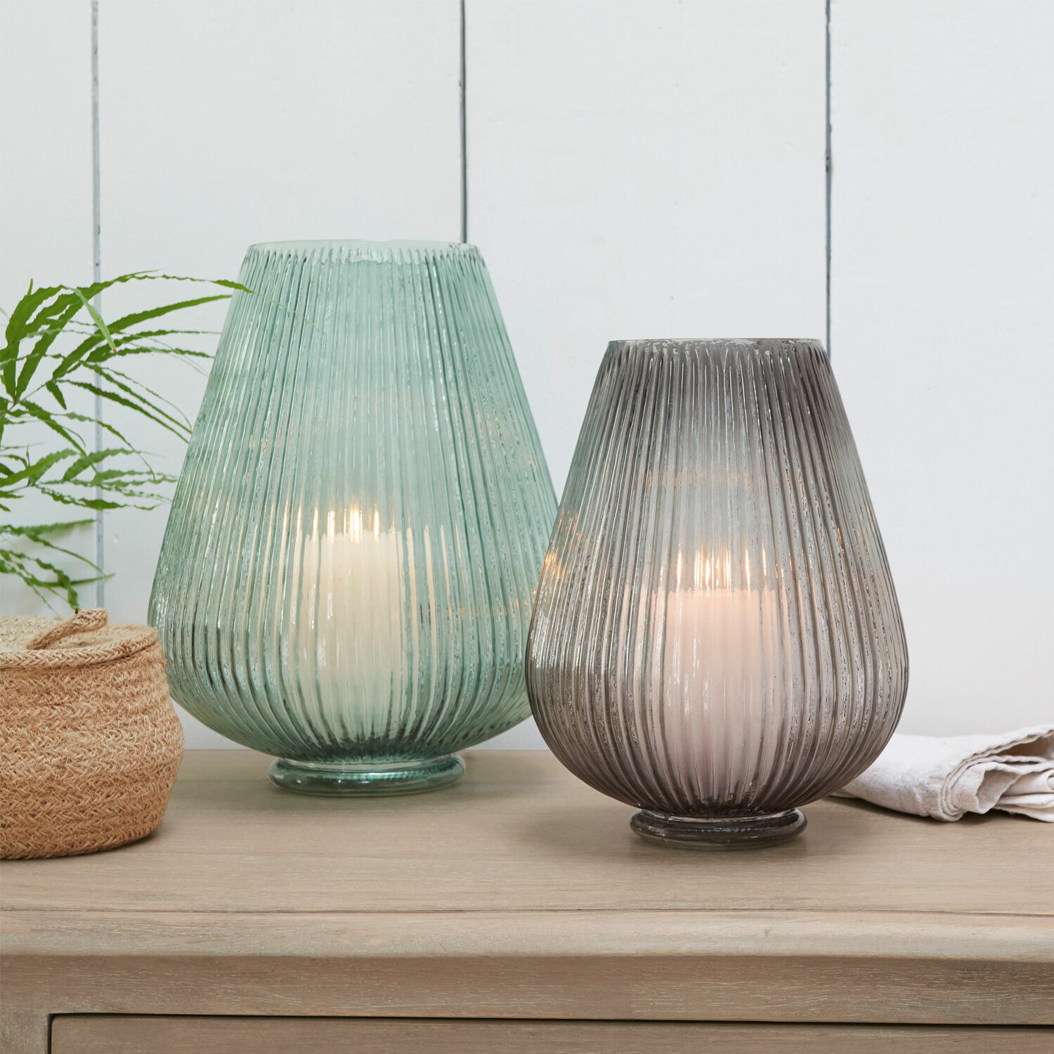 Twee glazen lantaarns in groen en grijs met witte kaarsen op een lichte houten tafel