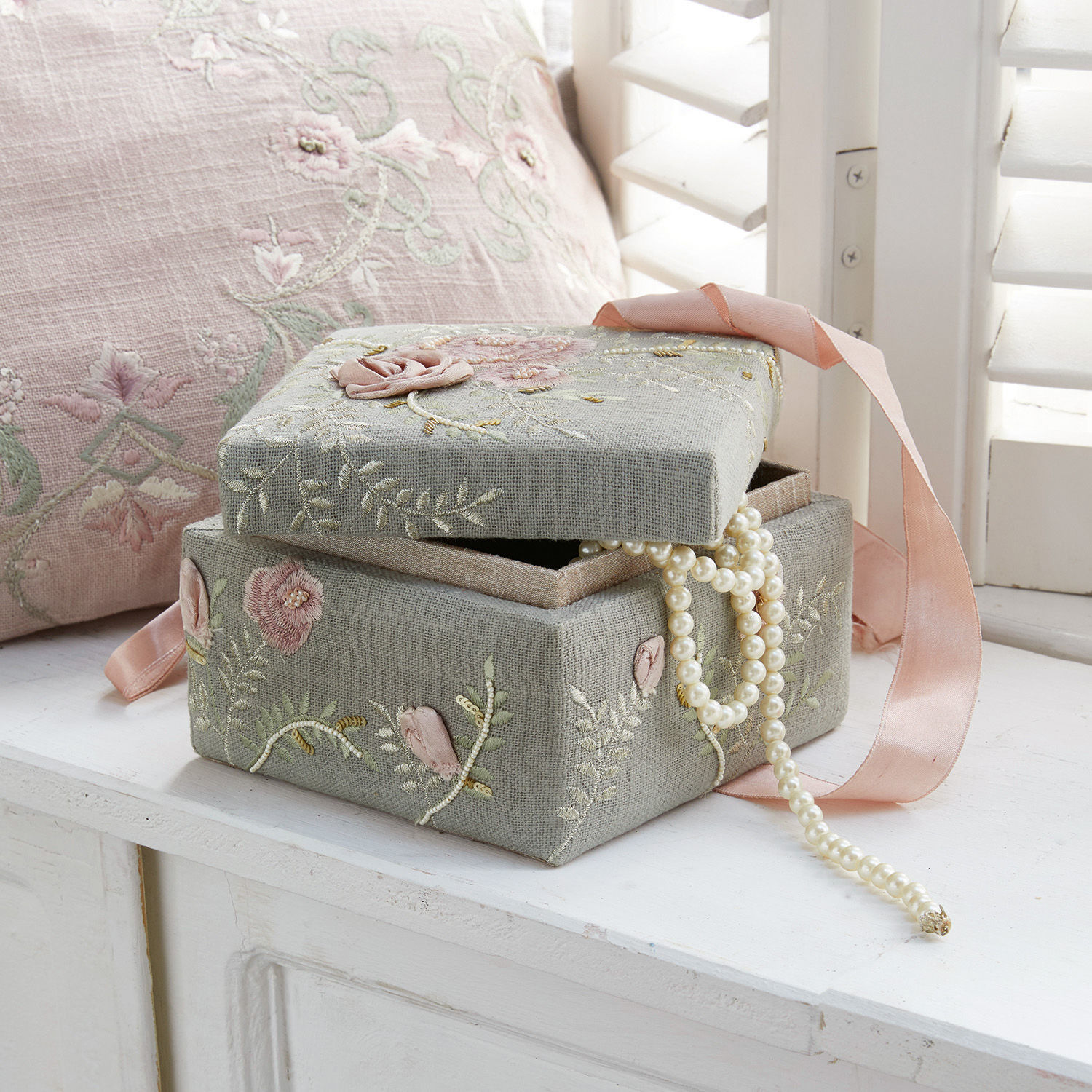 Grijze linnen sieradendoos met bloemenborduursel en roze lint, parelketting zichtbaar