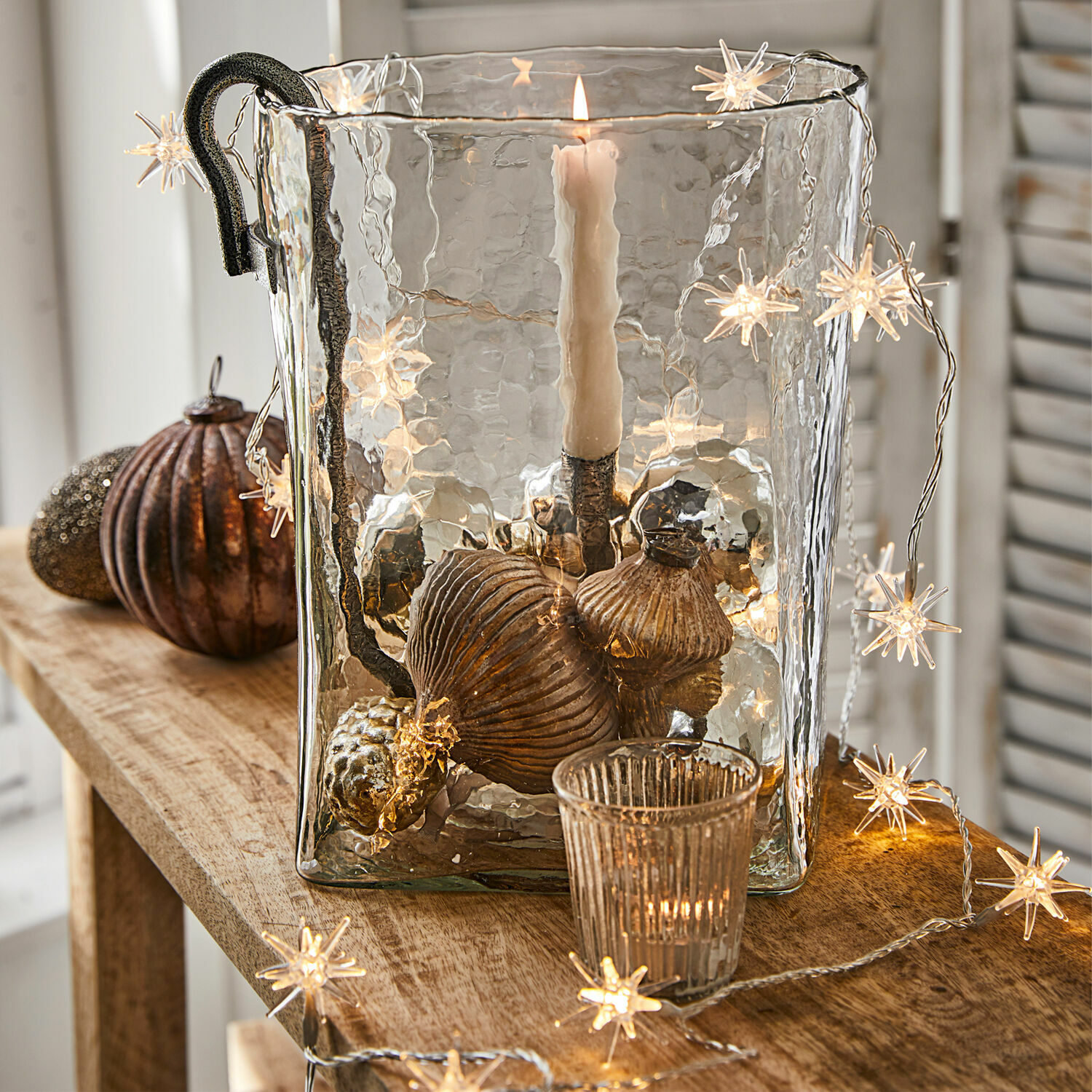 Lichtsnoer Nelisse Glazen windlicht met metalen handvat en koperen decoraties op houten tafel met stervormige lichtjes