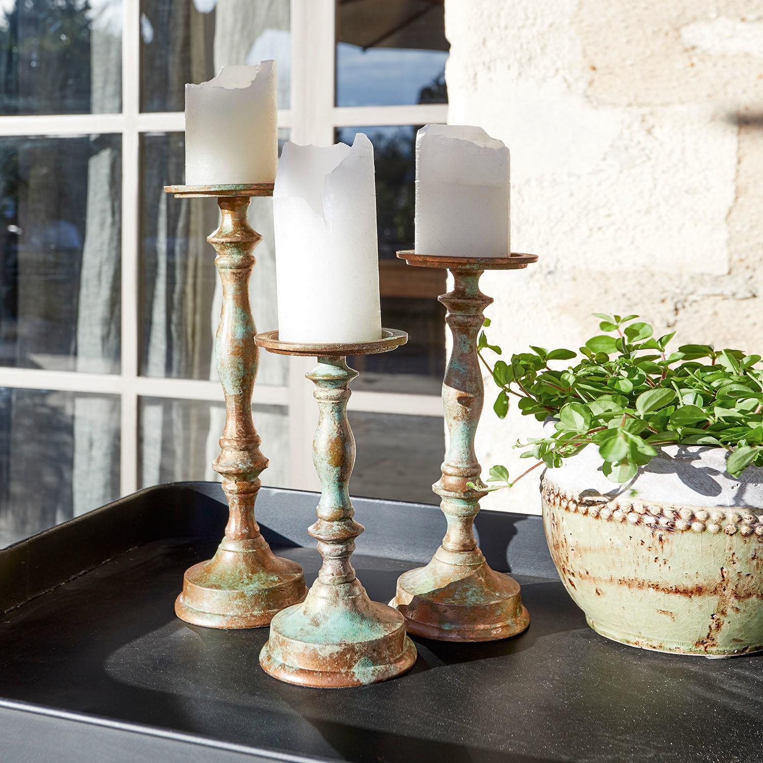 Drie messing kandelaren met deels gesmolten witte kaarsen op een zwart dienblad naast een pot met groene plant