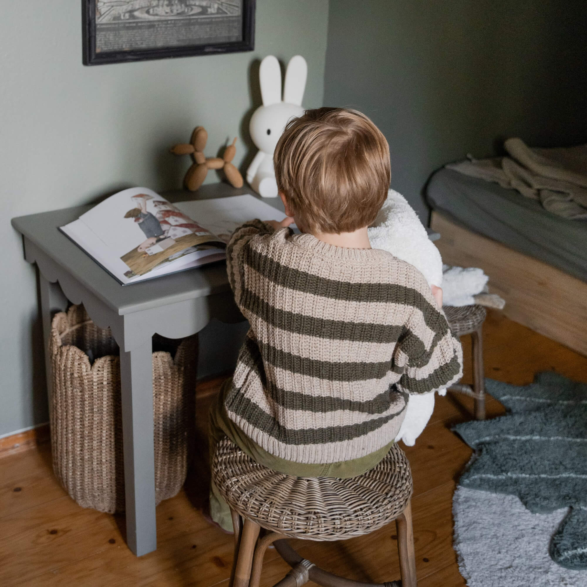 Kind in gestreepte trui zit op rotan kruk bij grijze tafel met boek en konijnenlamp