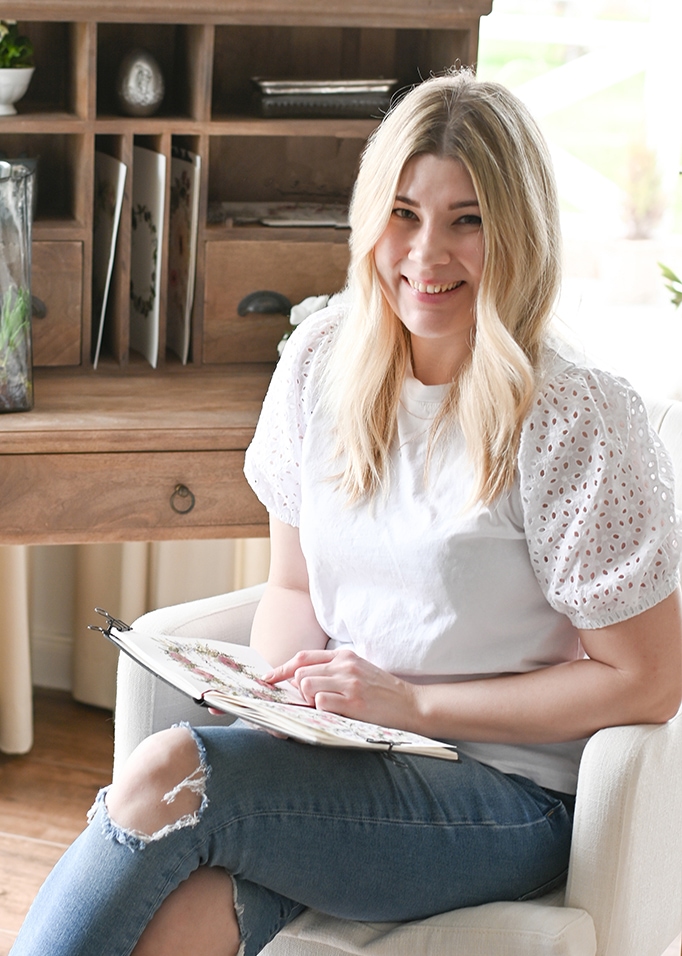 Jonge vrouw in witte blouse met opengewerkte mouwen en gescheurde spijkerbroek zit met album in lichte kamer