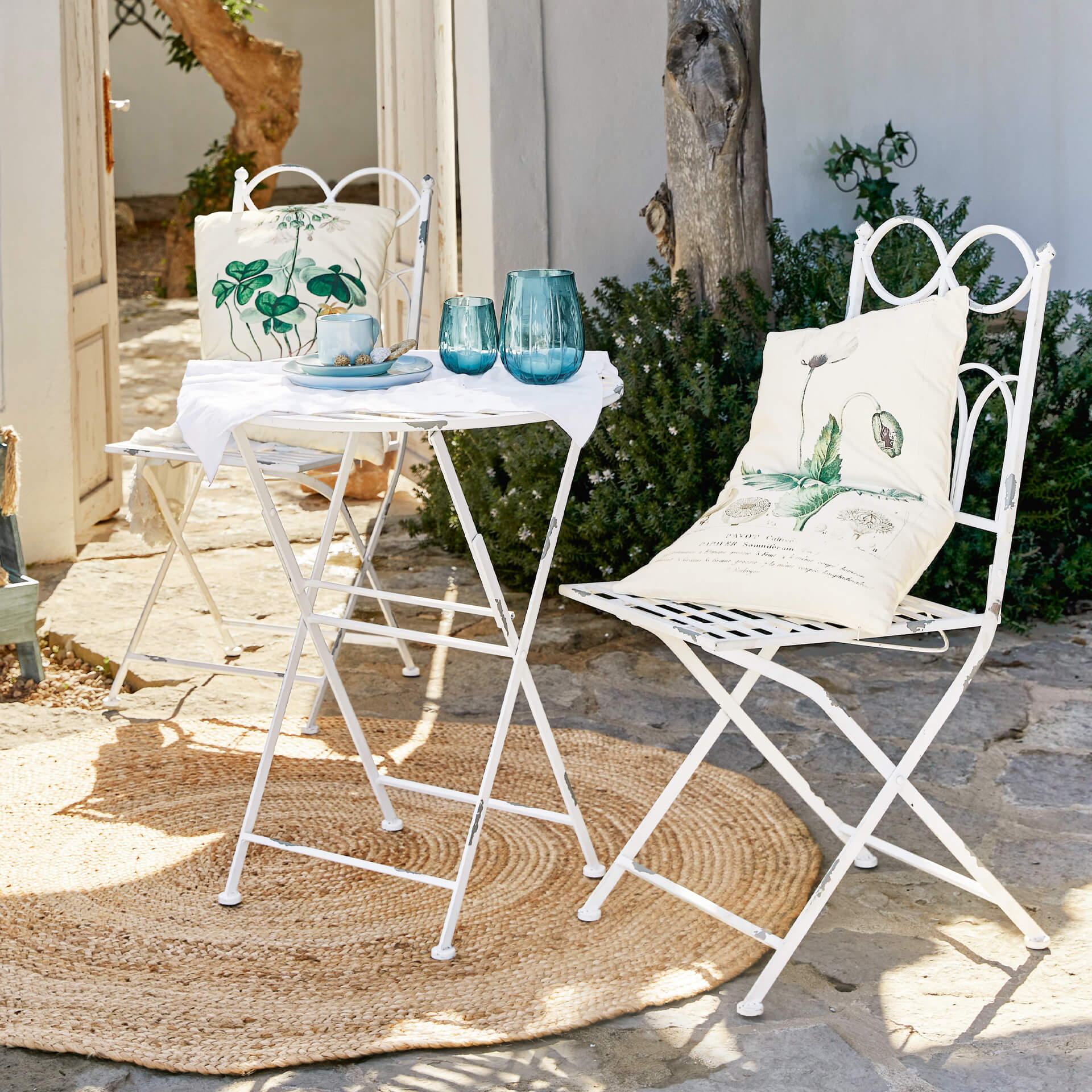 Witte metalen tuinset met kussens met groene bladeren op stenen terras met jute vloerkleed