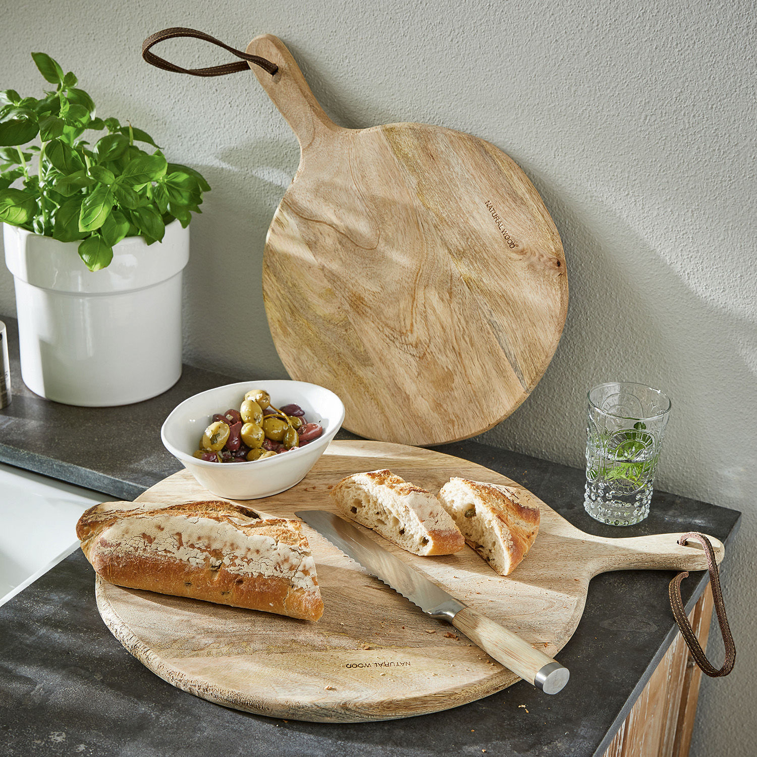Snijplank set van 2 Nitra Rond snijplank van natuurlijk hout met handvat, broodmes, gesneden brood en olijven op keukenblad