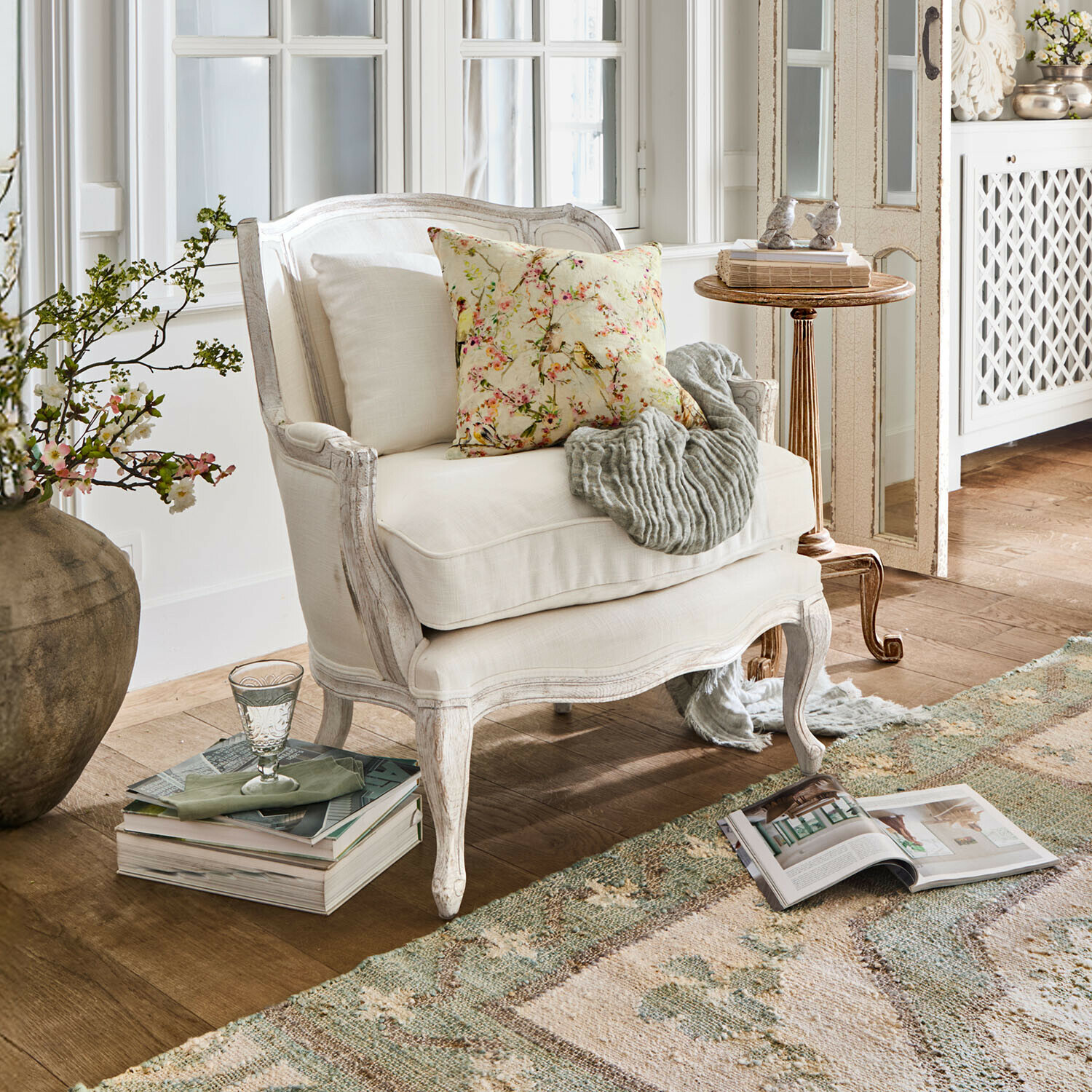 Witte houten fauteuil met lichte bekleding, bloemenkussen en grijze deken in gezellige woonkamer