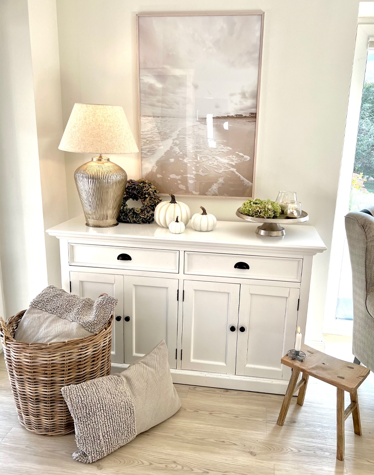 Witte houten dressoir met lamp, decoratieve pompoenen en rieten mand met kussens in lichte woonkamer