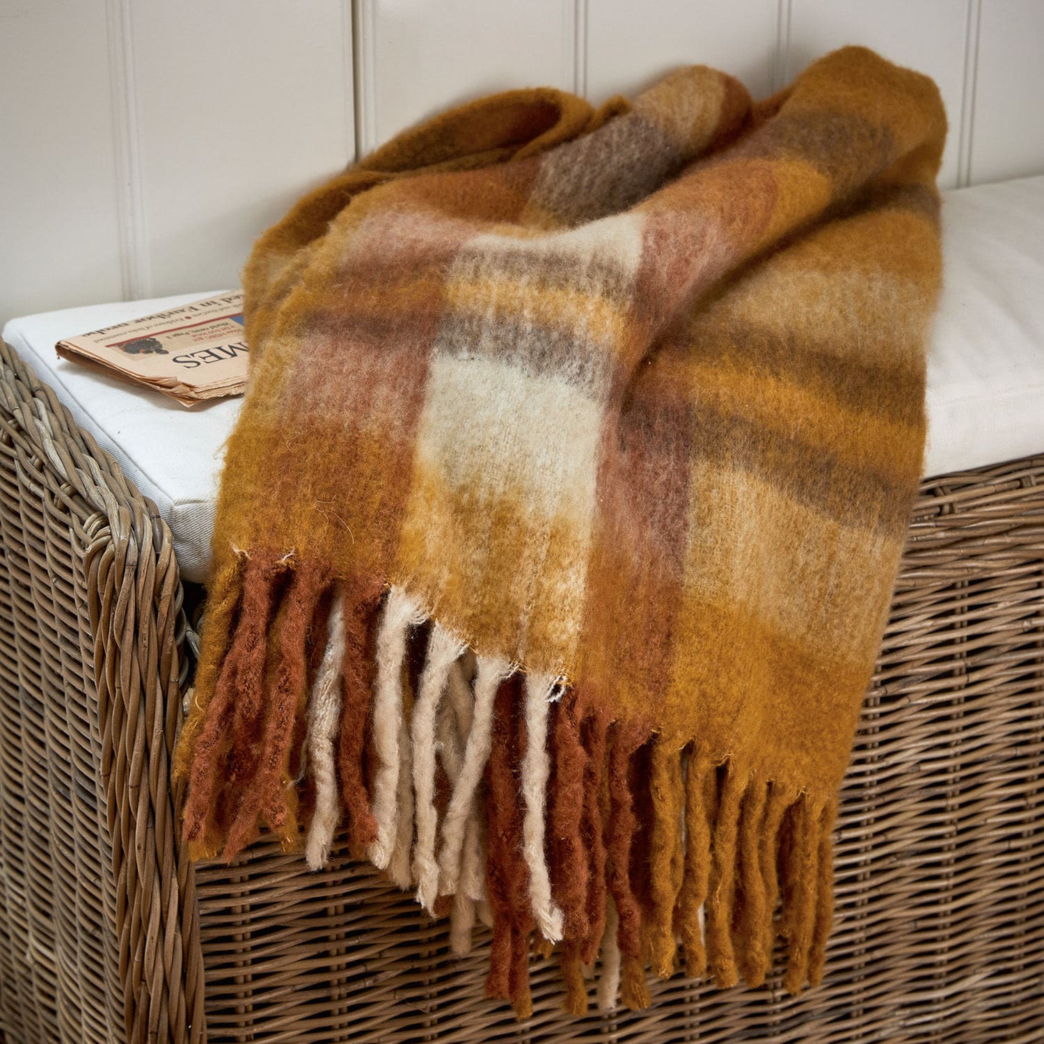 Plaid Alborg Zachte wollen plaid in warme bruin- en beigetinten met franjes op een rotan stoel