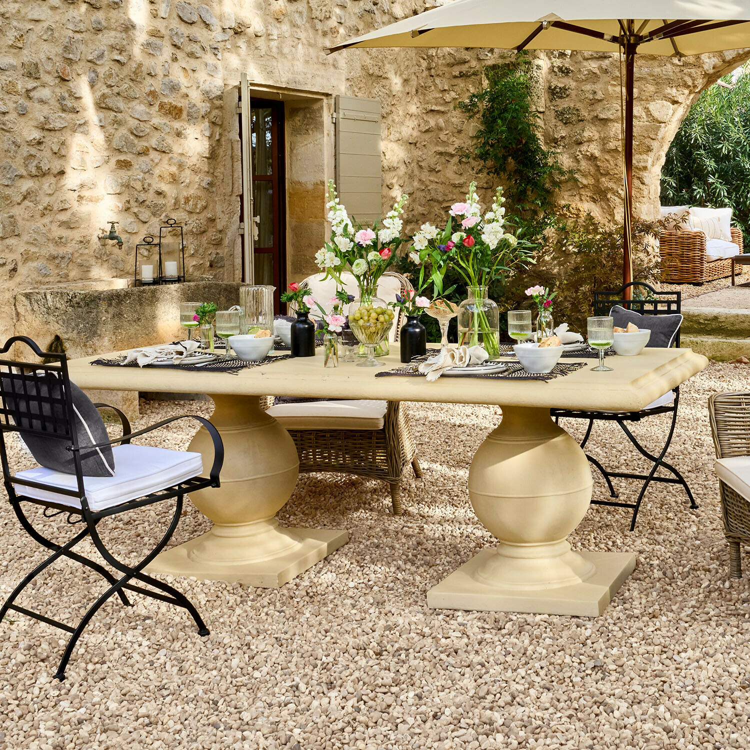 Grote beige stenen tuintafel op grindpatio met bloemen en metalen en rieten stoelen