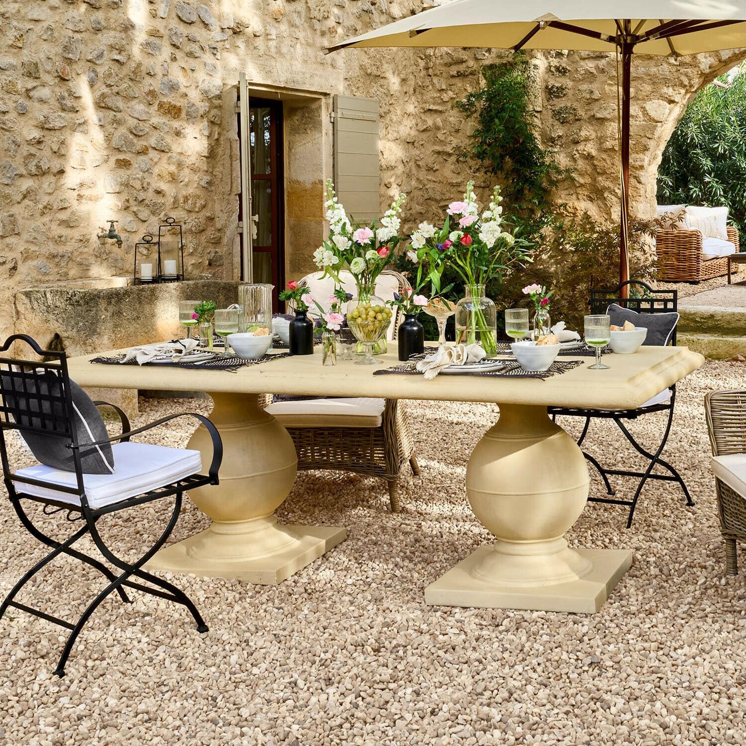 Tafel Talvero Grote beige stenen tuintafel op grindpatio met bloemen en metalen en rieten stoelen