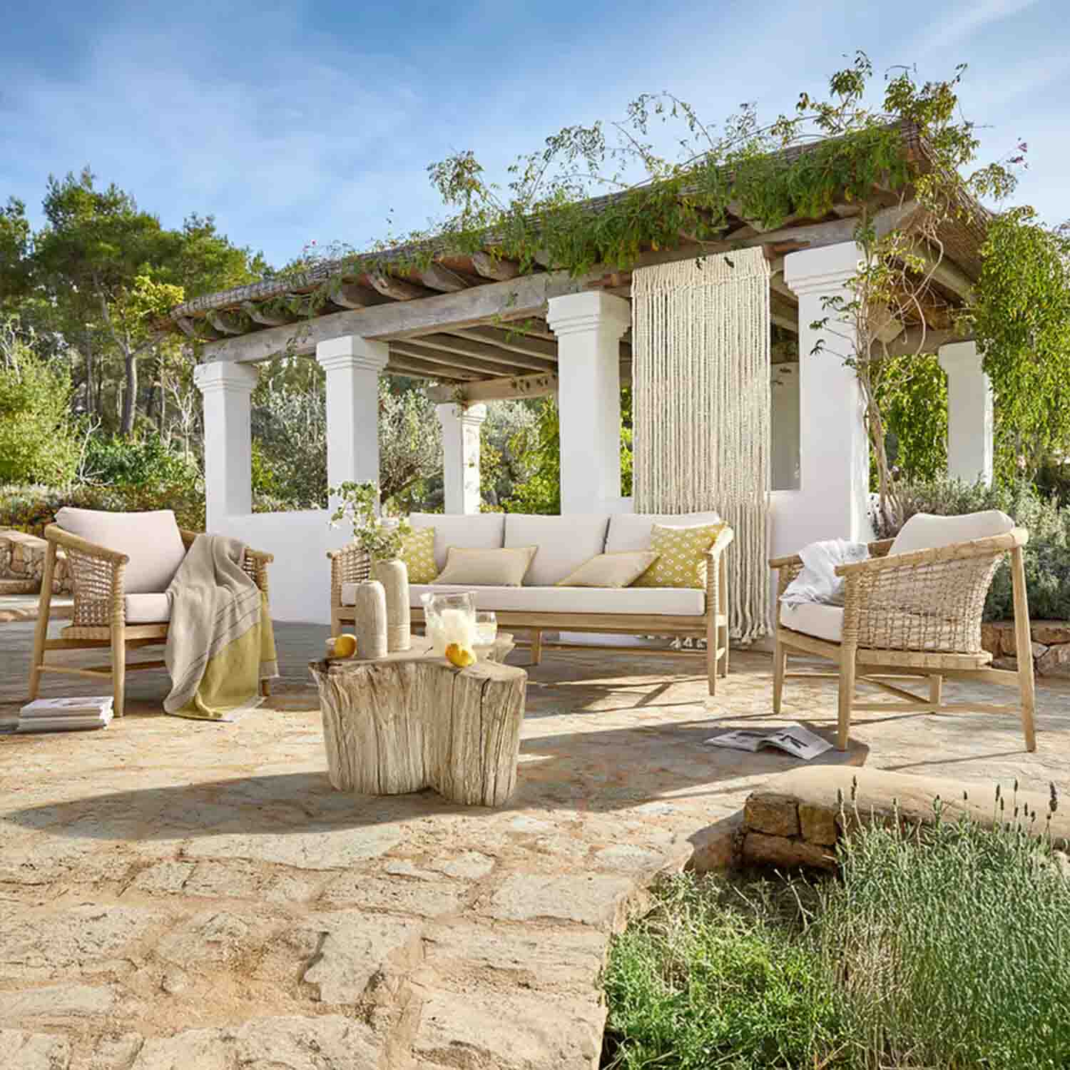 Tuinmeubelset van licht hout met crèmekleurige kussens op stenen terras onder een met planten begroeide pergola