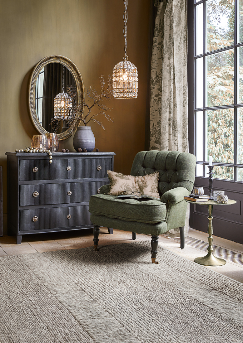 Gezellige groene fauteuil met kussens naast zwarte ladekast en gouden bijzettafel in woonkamer met vloerkleed