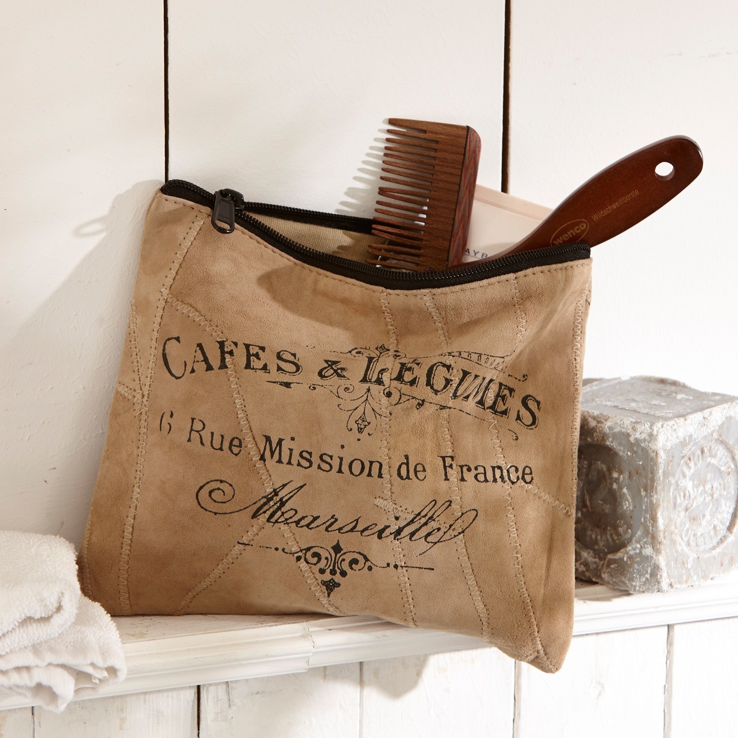 Beige suède make-uptasje met zwarte rits en Franse opdruk, houten kam zichtbaar op plank