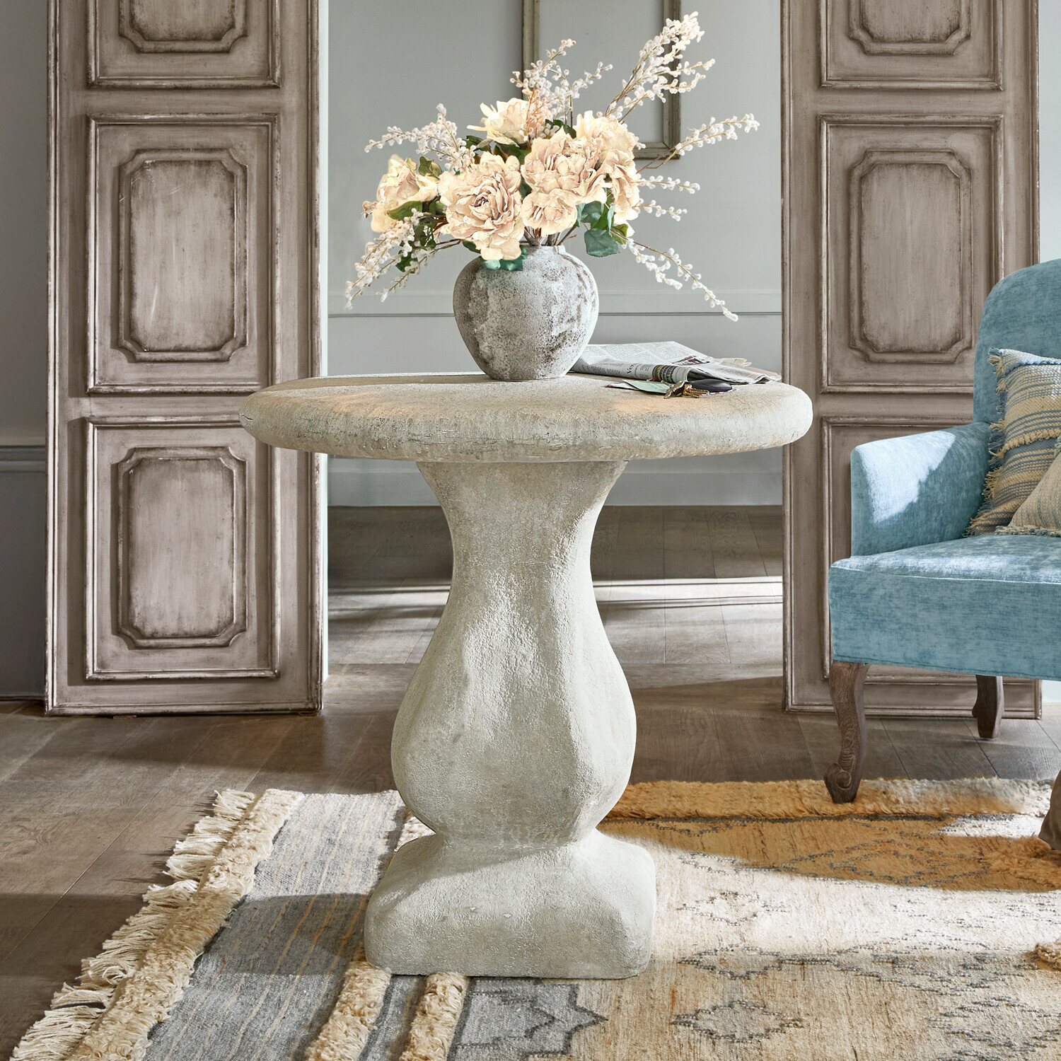 Ronde grijze betonnen tafel met vaas en bloemen in woonkamer met lichtblauwe stoel