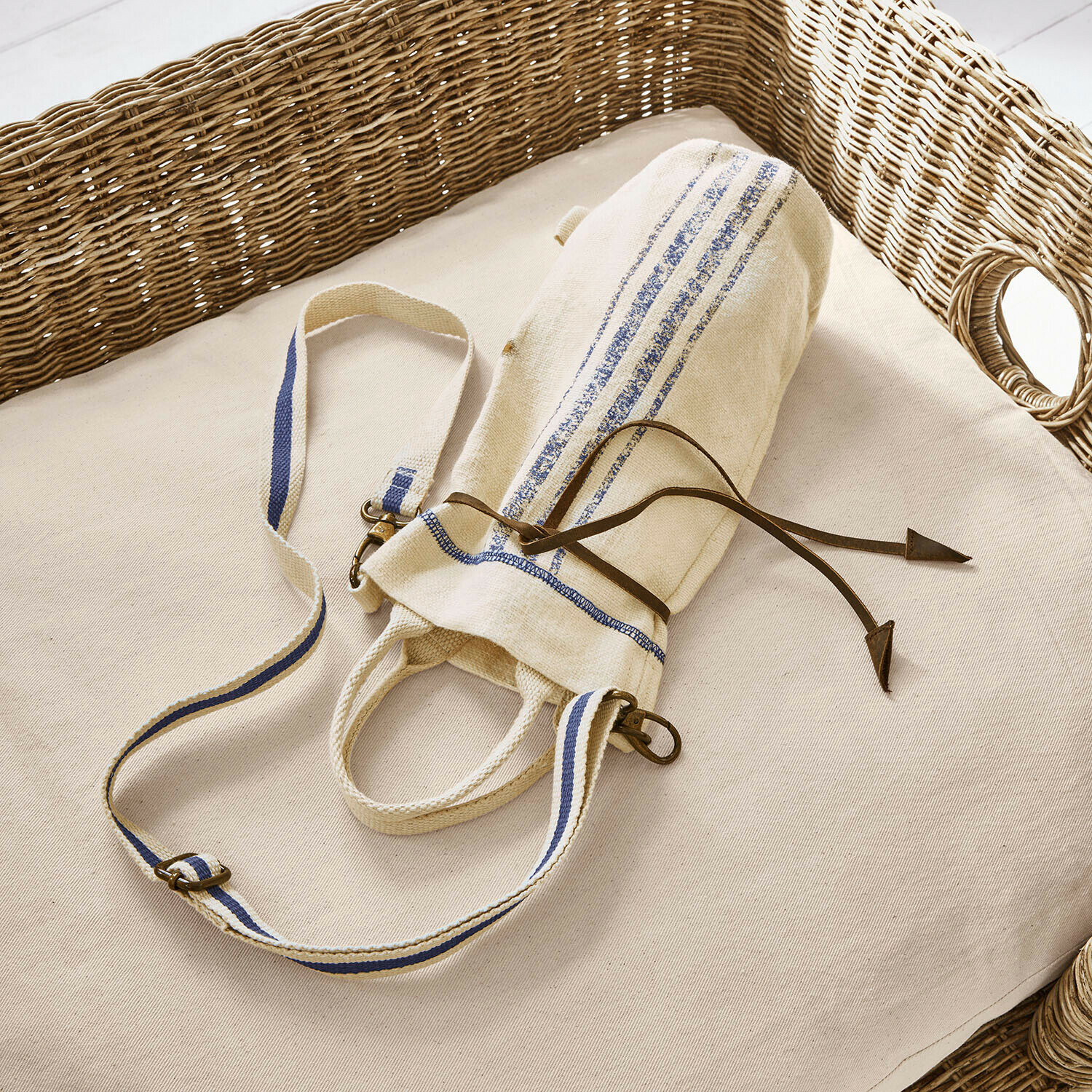 Flessentasje Pawera Beige katoenen tas met blauwe strepen en leren koord op licht kussen met rieten rugleuning