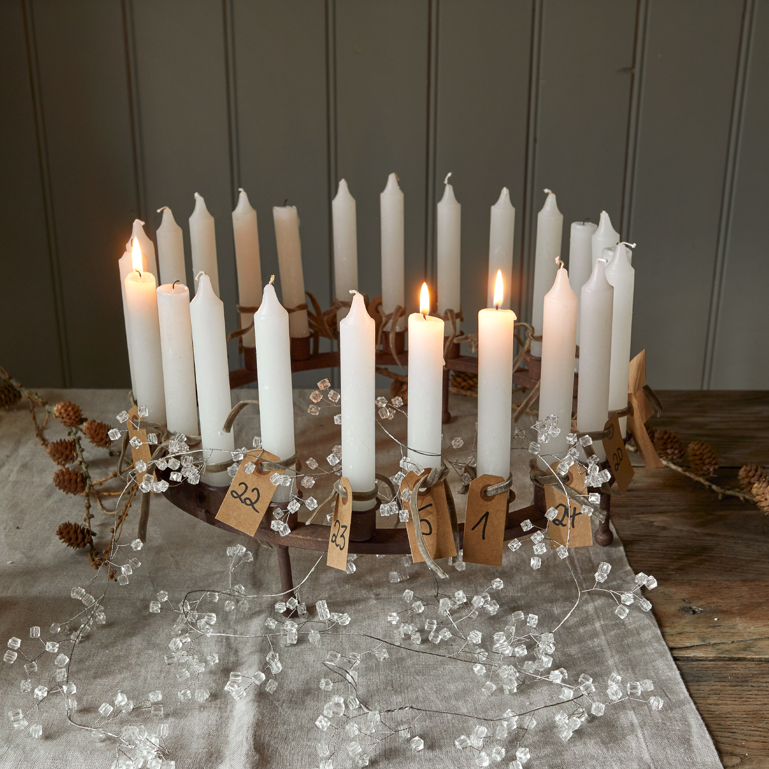 Metalen adventskrans met 24 witte kaarsen en genummerde papieren labels op linnen tafelkleed