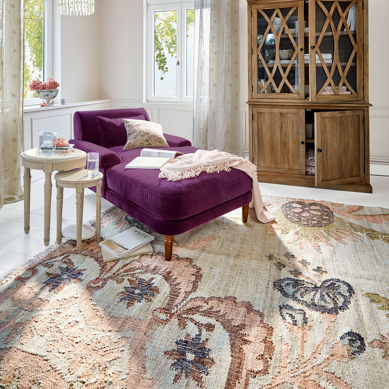 Paars fluwelen ligstoel in lichte woonkamer met houten vitrinekast en decoratief vloerkleed