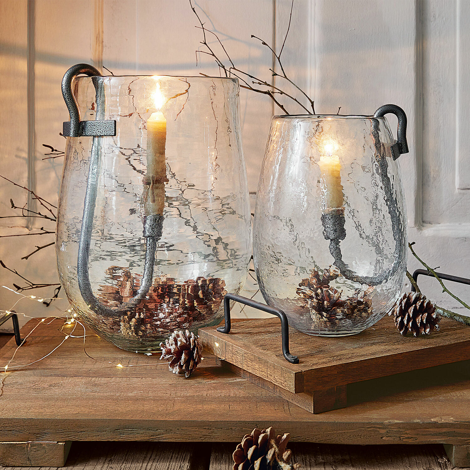 Windlicht set van 2 Lorcalla Twee transparante glazen lantaarns met metalen handvatten en kaarsen, versierd met dennenappels