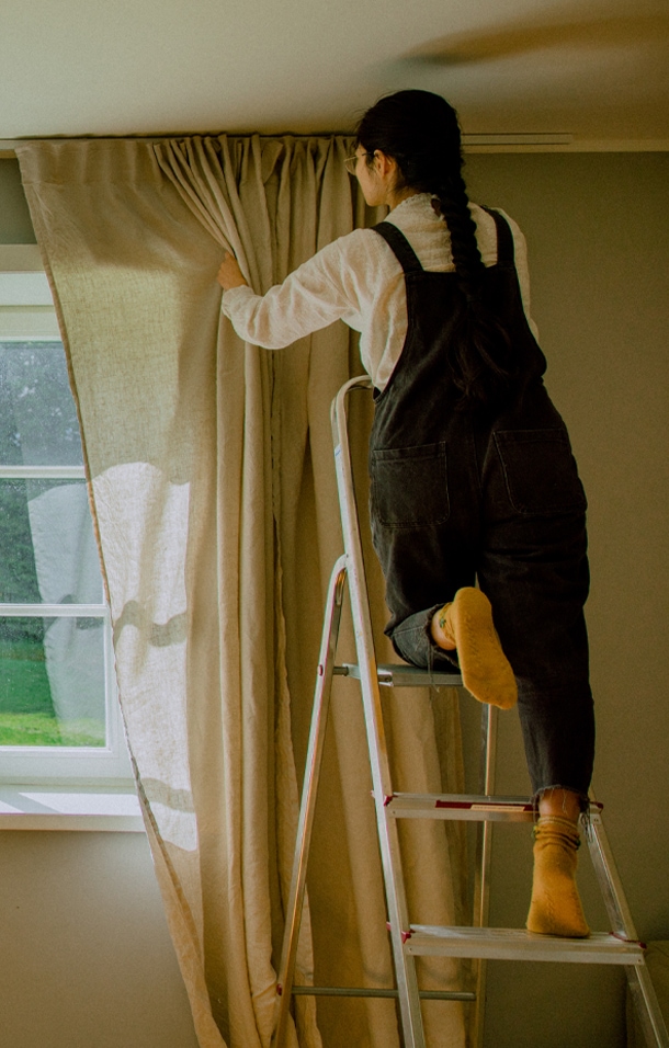 Vrouw op ladder die beige linnen gordijn voor raam in lichte kamer sluit