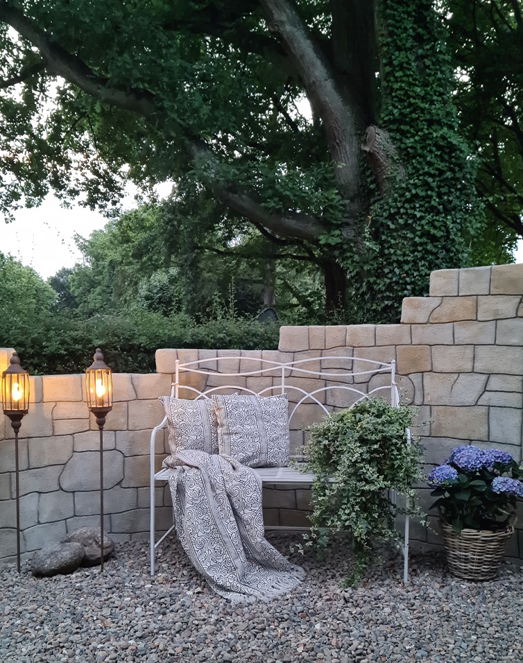 Witte metalen tuinbank met kussens en deken, omgeven door stenen muur en planten
