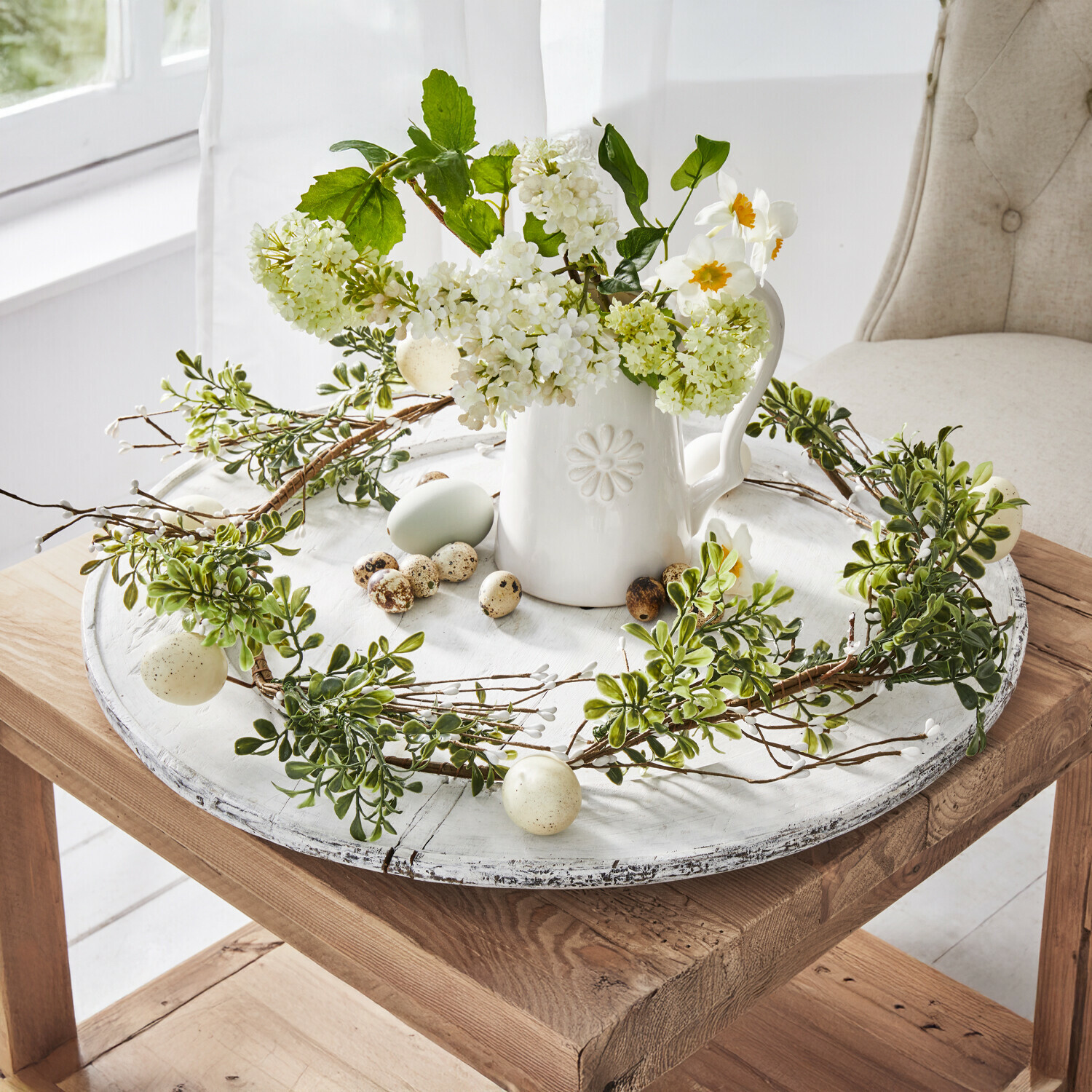 Decoratieve krans van groene bladeren en eieren op een wit houten dienblad met keramische kan en bloemen