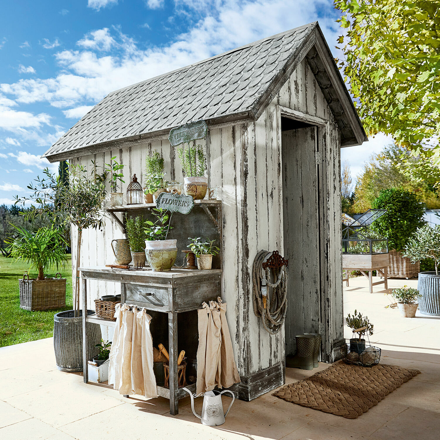 Witte houten tuinberging met verweerde look en metalen plantenbank op zonnig terras