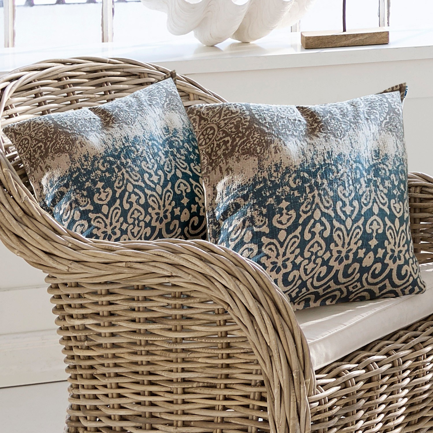 Kussen Rod Twee decoratieve kussens met blauw-beige patroon op een natuurlijke rotan stoel