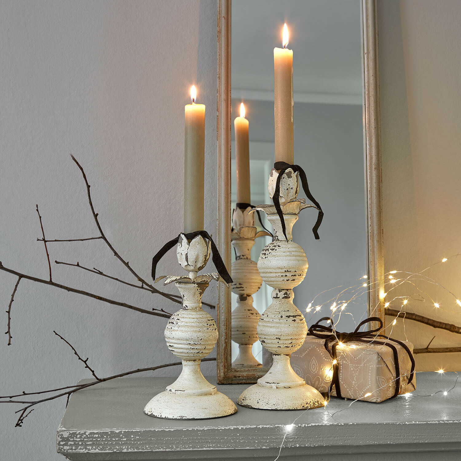 Twee crèmekleurige metalen kandelaars met beige kaarsen en zwart lint op grijze plank met lichtsnoer
