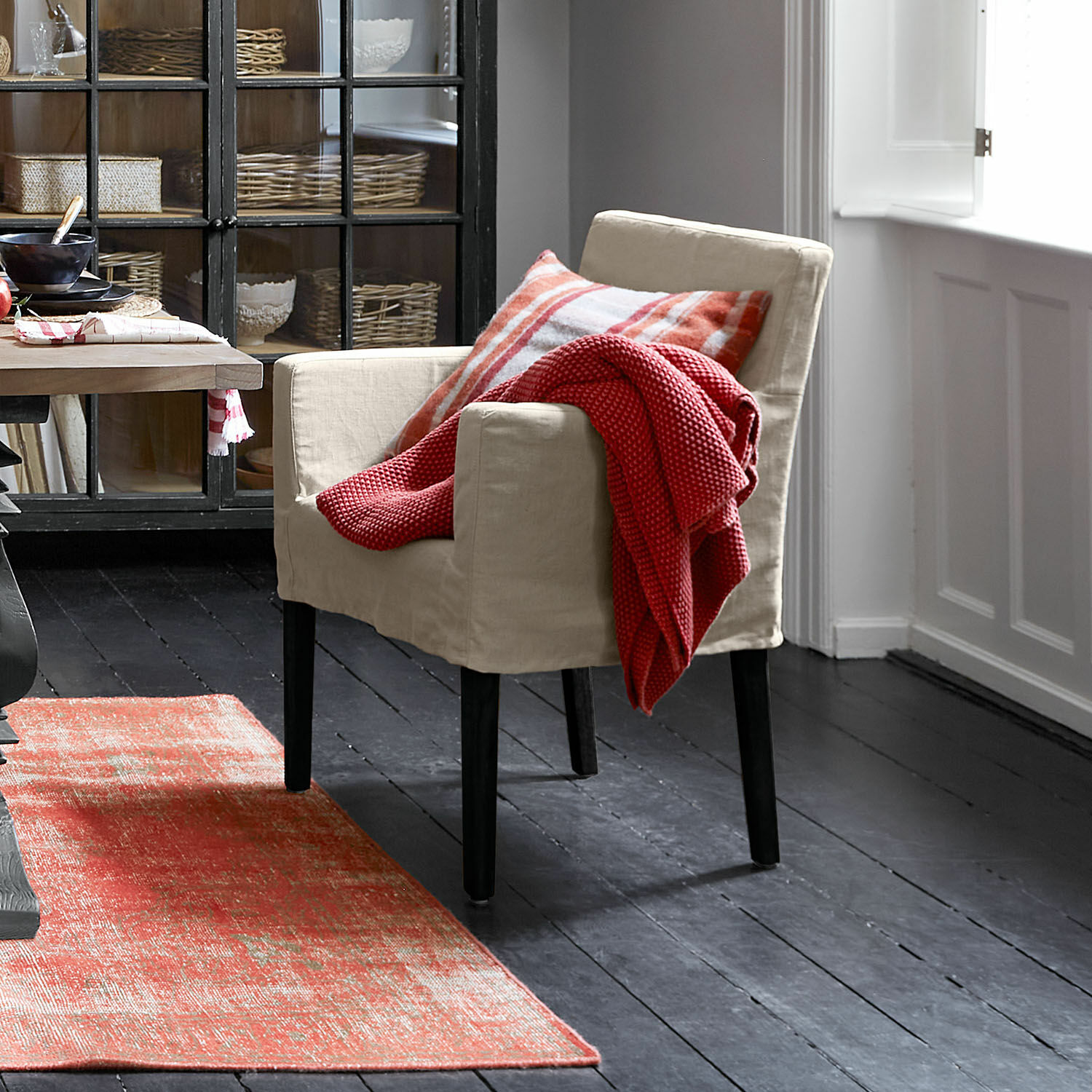 Beige stoffen fauteuil met zwarte poten, rode deken en geruit kussen, rode vloerkleed