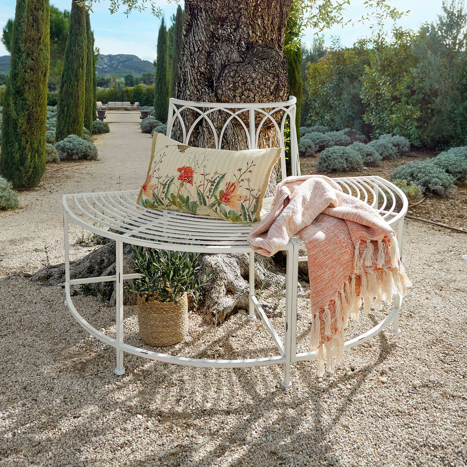 Witte metalen tuinbank rond een boom met bloemenkussen en oranje deken op grindpad