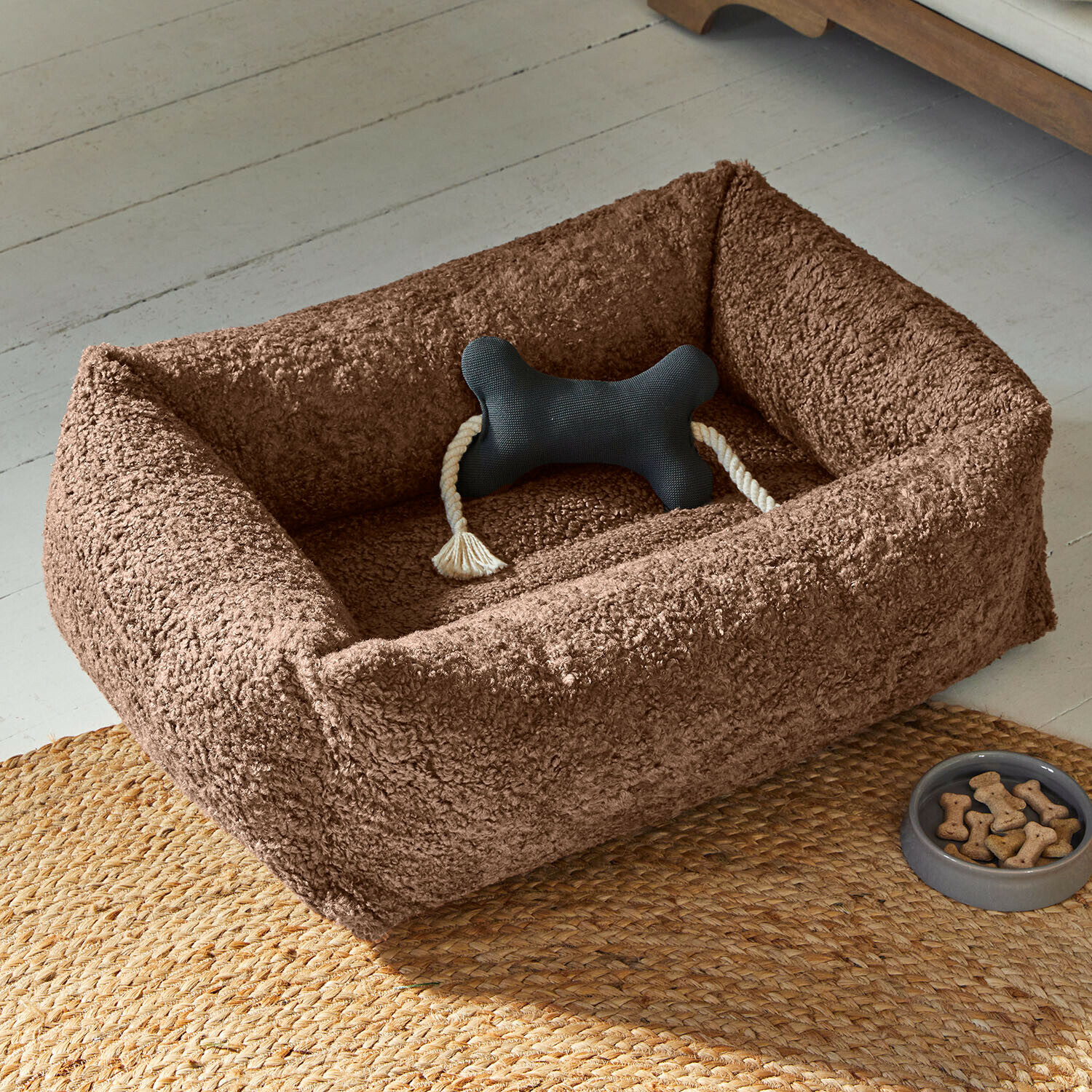 Bruin pluche hondenmand met zwart bot-speelgoed en bakje met traktaties op gevlochten kleed