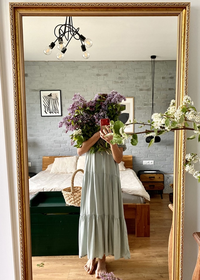 Vrouw in lange groene jurk met paarse bloemenboeket voor spiegel in slaapkamer met houten bed