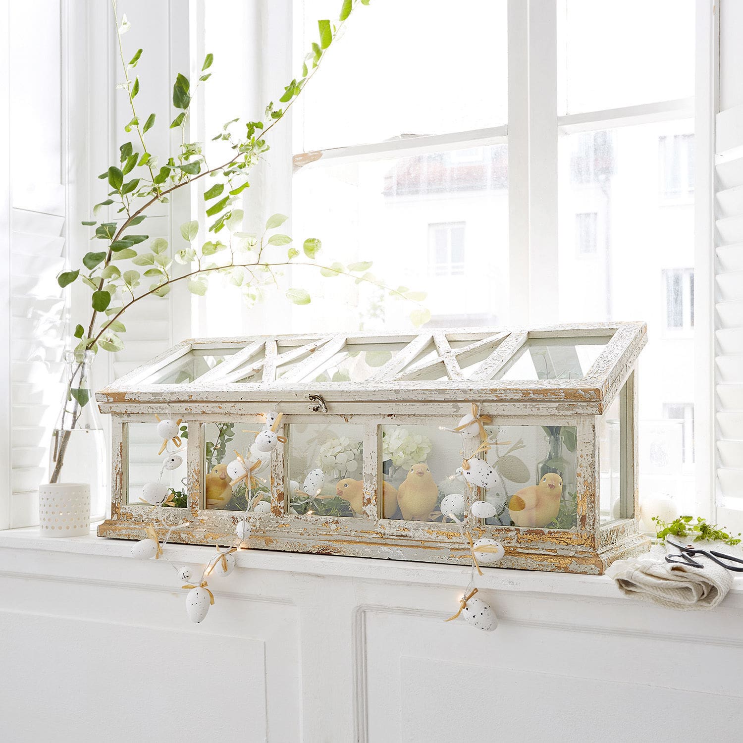 Witte houten mini-kas met verweerde look en kuikendecoratie op een vensterbank