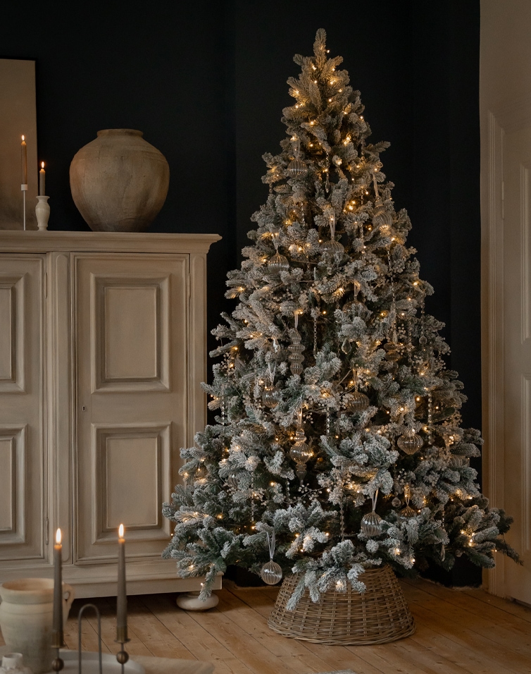 Kunstmatige besneeuwde kerstboom met warme verlichting en glazen versieringen in rieten standaard