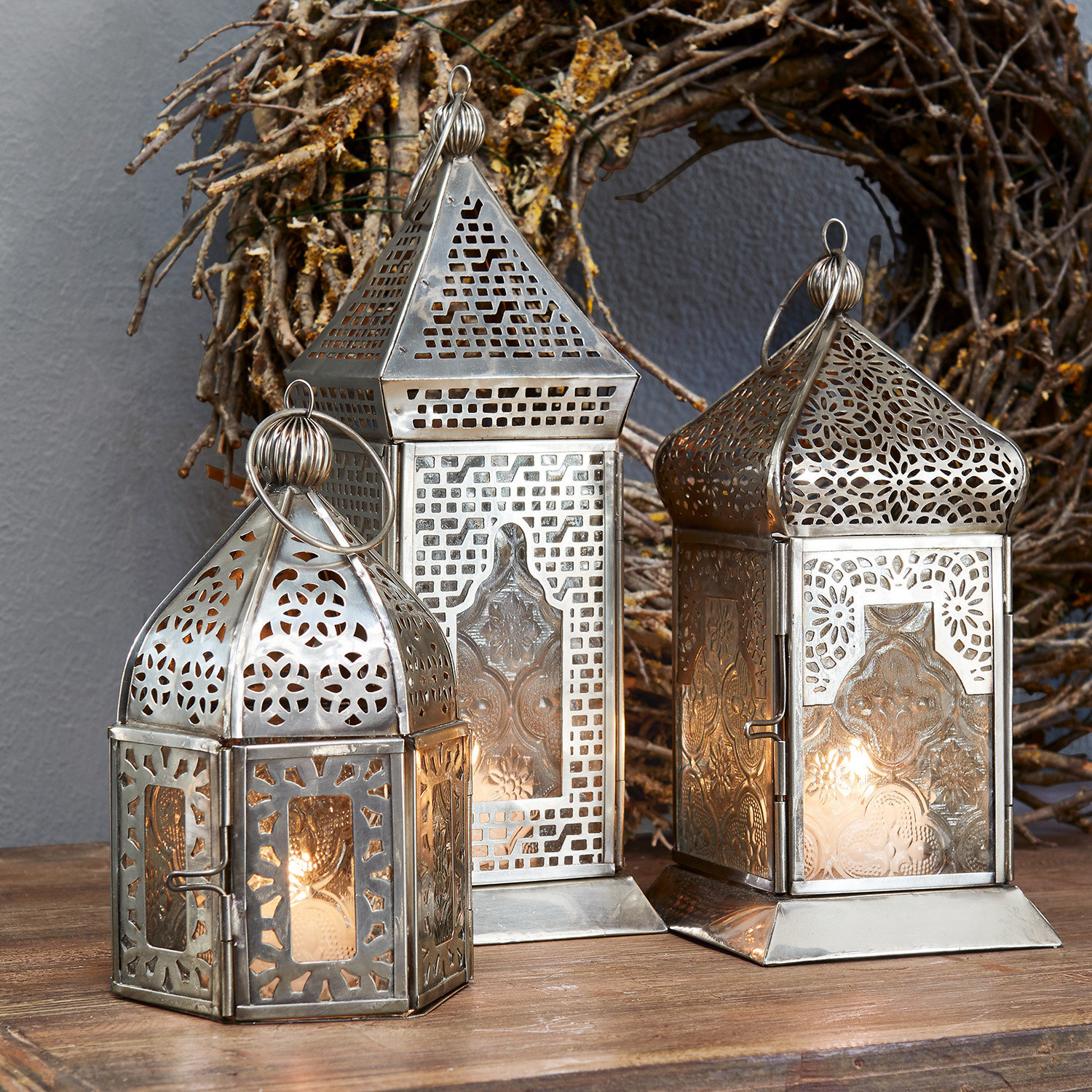 Drie zilverkleurige metalen lantaarns met opengewerkte patronen en glazen panelen op houten tafel